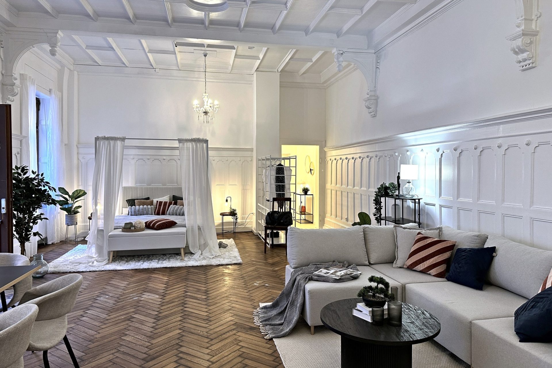 Home Staging in einem denkmalgeschützten Tanzsaal in Karlsruhe: helles Sofa im Vordergrund, dahinter ein Bett mit leichten Vorhängen, Stuckdecke und weiße Vertäfelung, Fischgrätparkett.